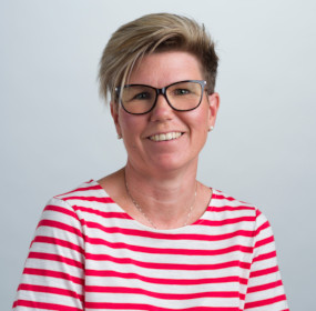 Leende person med kort, ljusbrunt hår med glasögon och en röd-vitrandig skjorta, mot en vanlig bakgrund.