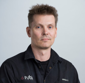 En person med kort hår klädd i en skjorta med mörk krage står mot en vanlig ljus bakgrund.