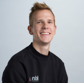 Person med kort hår klädd i en svart Rubi-tröja och ler mot en ljus bakgrund.