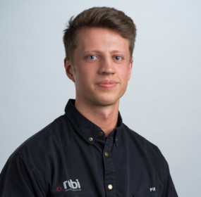 Person med kort hår klädd i en svart skjorta med "rbi" och initialer broderade på mot en vanlig bakgrund.