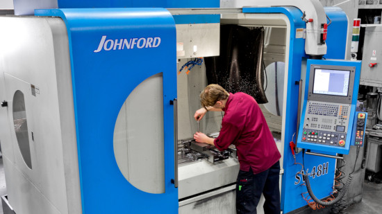 Person som använder en blå Johnford ST-40H CNC-maskin, justerar komponenter inuti maskinen med kontrollpanelen synlig till höger.